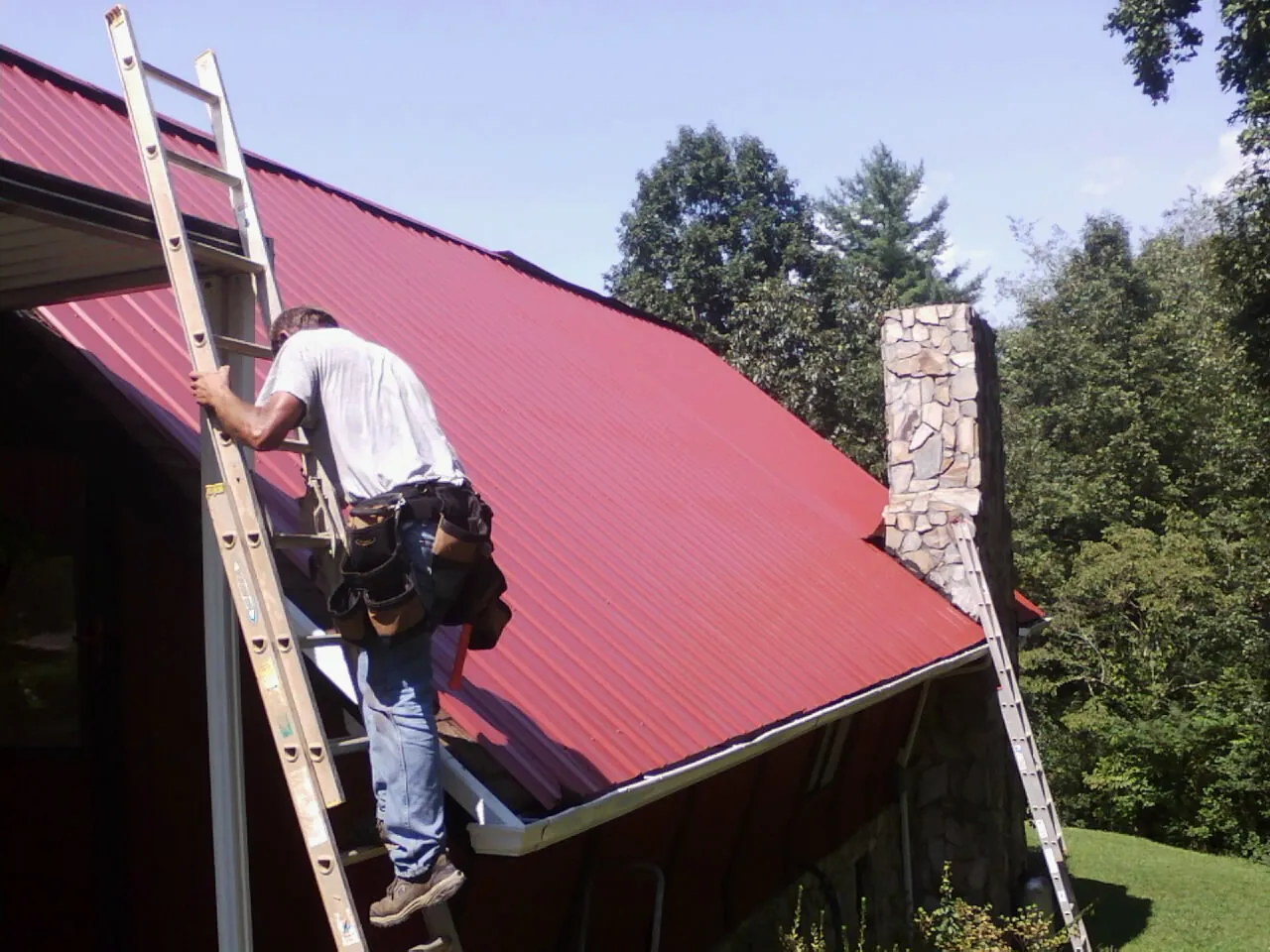 Roofer on a ladder beside a red metal roof and stone chimney — Soffit & Fascia Repair by Roofing Brothers in Laurel Springs